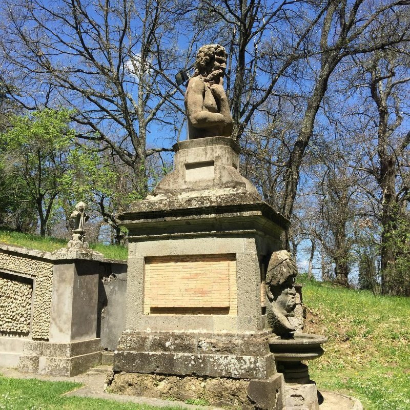 Auto-generated description: A stone monument set in a grassy area features a bust atop a pedestal, surrounded by trees and additional stone sculptures.