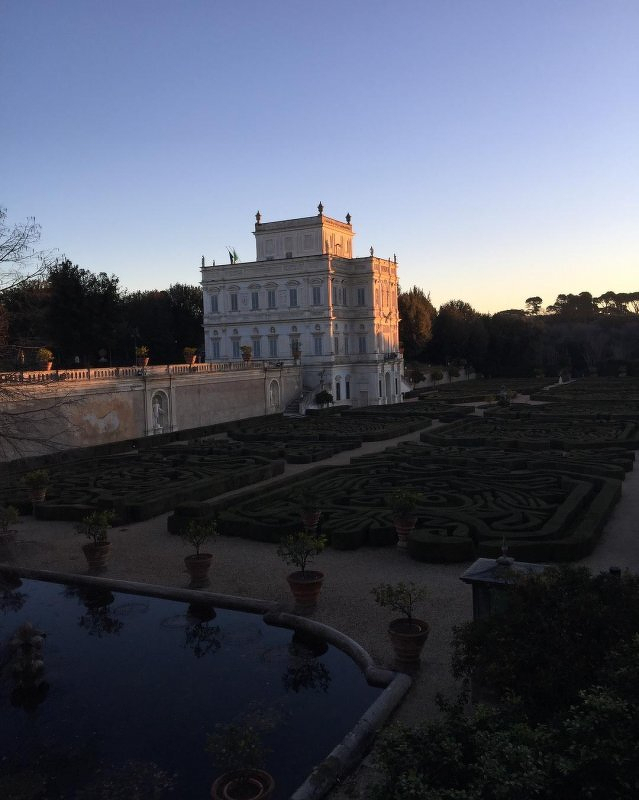 Auto-generated description: A historic villa is surrounded by formal, manicured gardens and a reflecting pool under a clear evening sky.