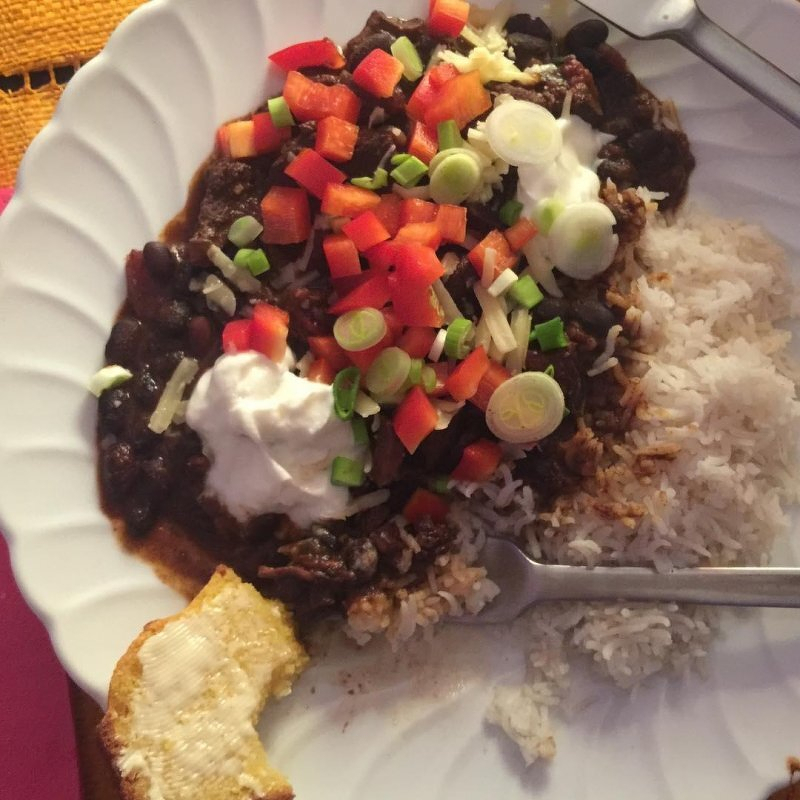 Auto-generated description: A plate featuring rice topped with a savory bean and vegetable mixture, garnished with diced red pepper, green onions, and dollops of sour cream, alongside a piece of bread.
