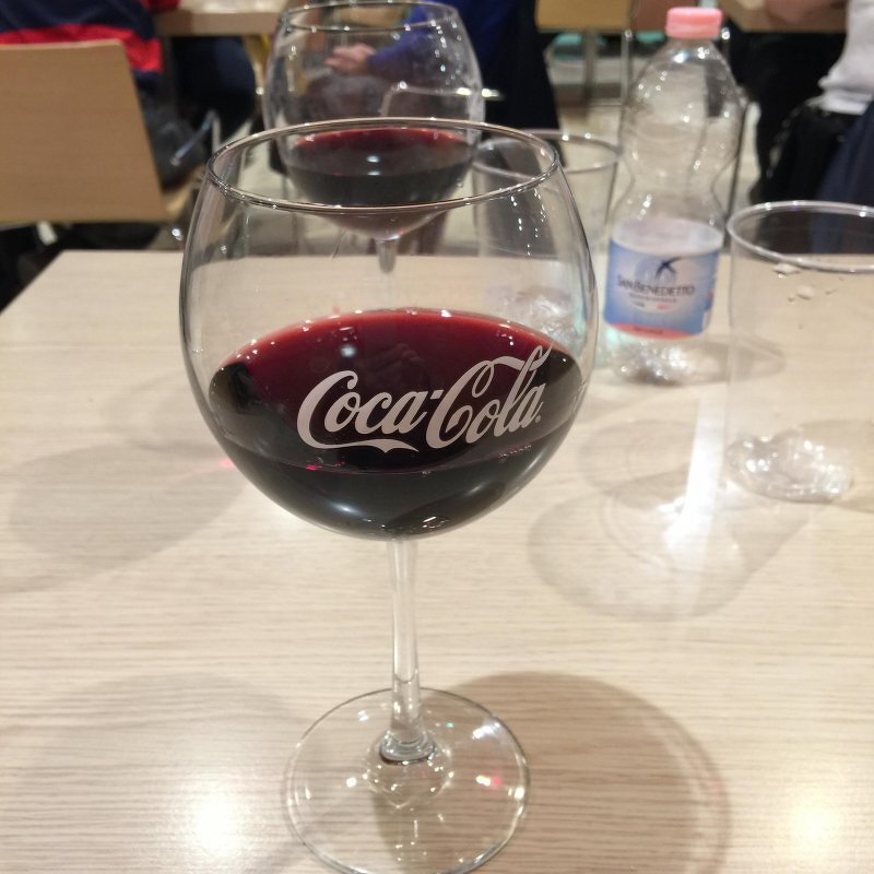Auto-generated description: A wine glass, unexpectedly featuring Coca-Cola branding, is filled with a dark liquid and placed on a table near a bottle of water.