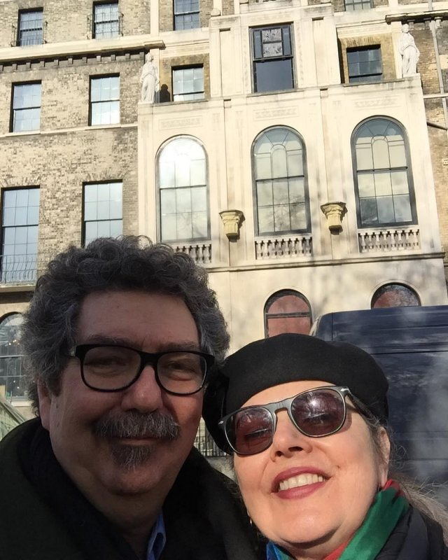 Auto-generated description: A smiling man and woman wearing glasses stand outdoors in front of a historic building with large windows and stone statues.