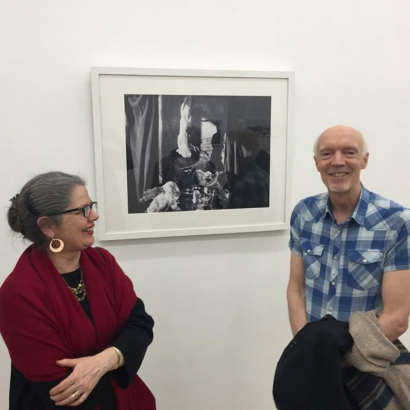Auto-generated description: A man and a woman are standing next to each other in a gallery, smiling at each other, with a framed black and white photograph on the wall behind them.