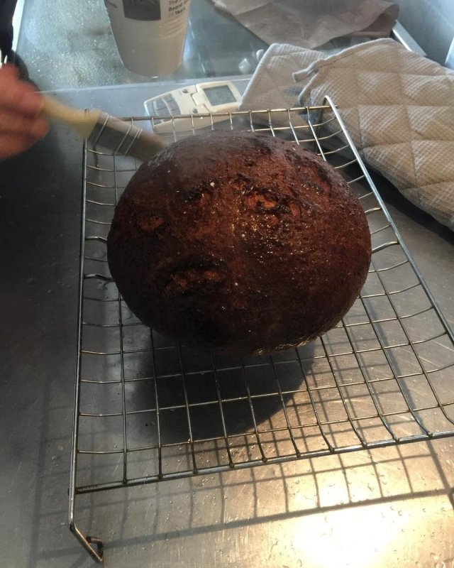 Auto-generated description: A hand is brushing glaze onto a round, freshly baked loaf of bread on a wire rack.