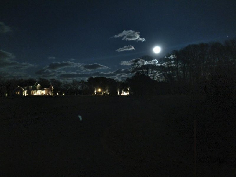 Auto-generated description: A bright full moon illuminates a dark landscape with clouds and distant house lights.