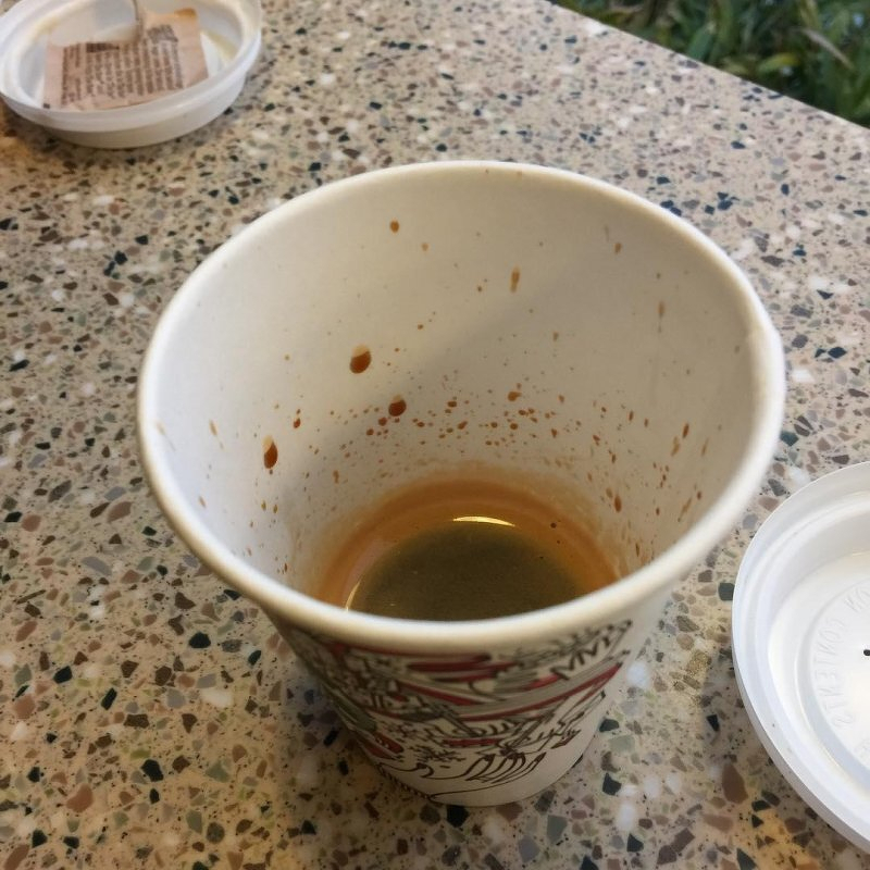 Auto-generated description: A nearly empty paper cup with some coffee residue is placed on a speckled surface, accompanied by plastic lids nearby.