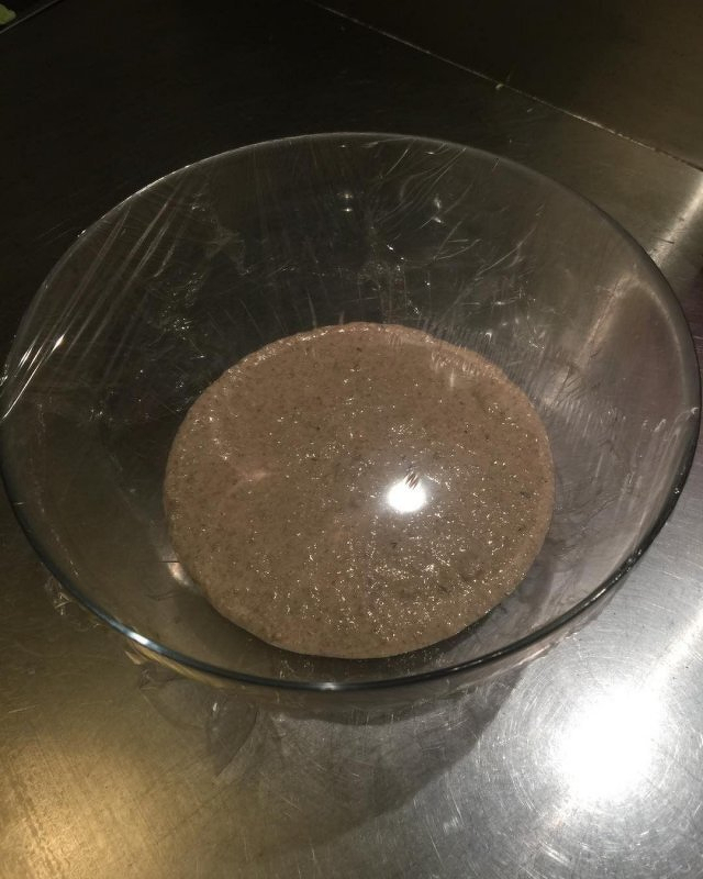 A glass bowl containing a dark brown mush and covered with plastic film is placed on a steel counter.