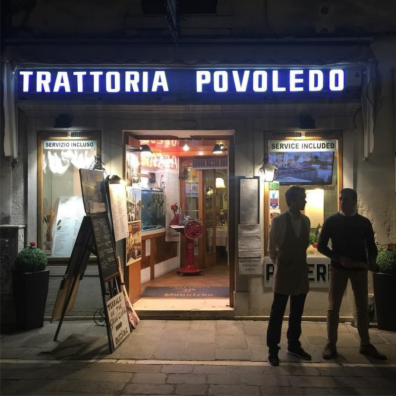 Auto-generated description: Two people stand in front of a well-lit restaurant entrance with the sign Trattoria Povoledo and various menus displayed.