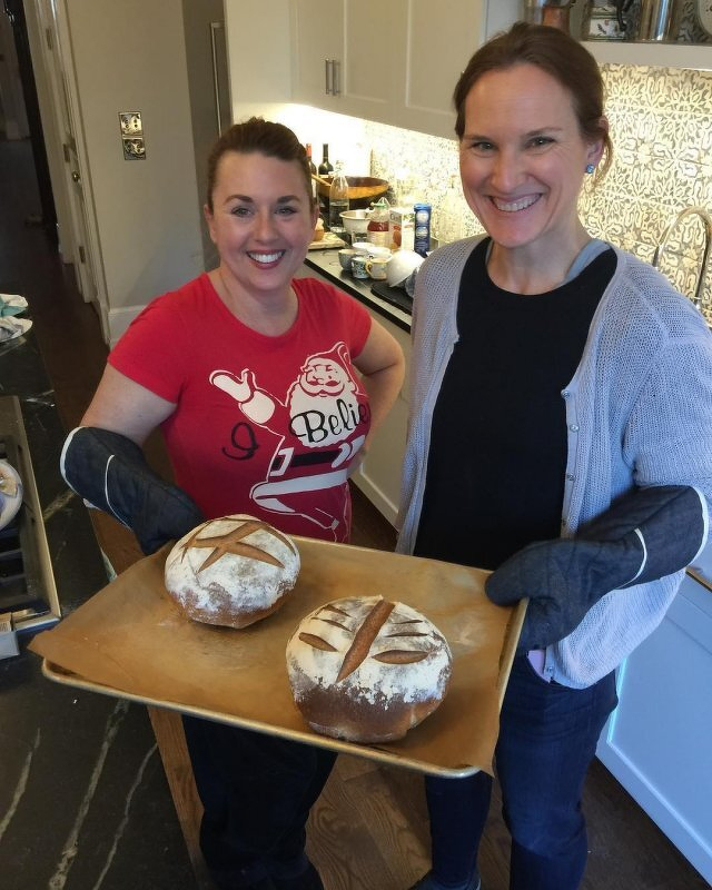 Two women hold a bread board on which are the two loaves they just pulled out of the oven.