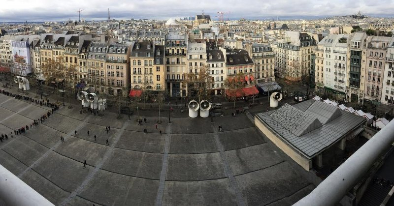 Auto-generated description: A wide cityscape view captures historic buildings lining a street with a large open plaza and modern architectural structures in the foreground under a cloudy sky. Human being adds: From the roof of the Centre Pompidou in Paris