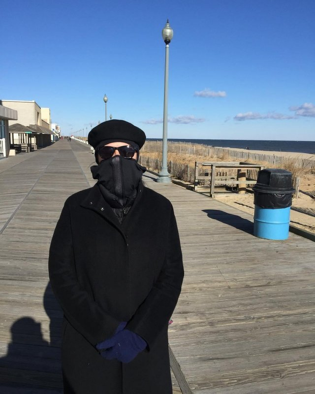 Auto-generated description: A person bundled in warm clothing stands on a boardwalk near the beach, under a clear blue sky.