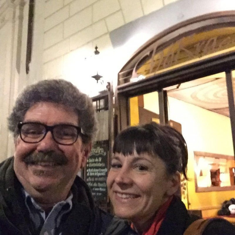 Auto-generated description: A man and a woman are smiling in front of a warmly lit building entrance.