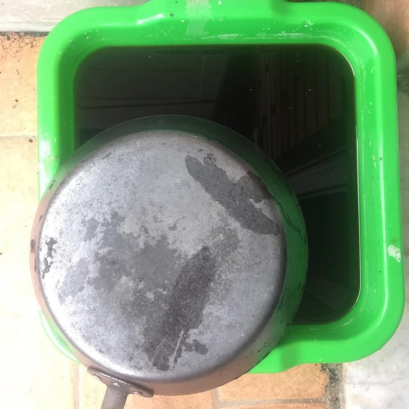 Auto-generated description: The underside of a cast-iron frying pan being restored, placed above a green square container filled with liquid.