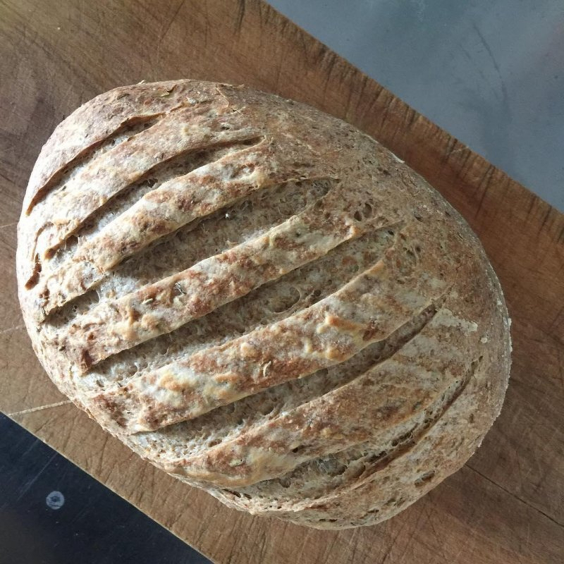 Auto-generated description: A crusty, round loaf of sourdough bread with a series of diagonal slashes on top rests on a wooden cutting board.