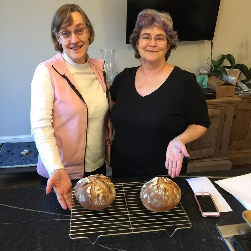 Two women gesture proudly at the loaves of bread they have just baked.