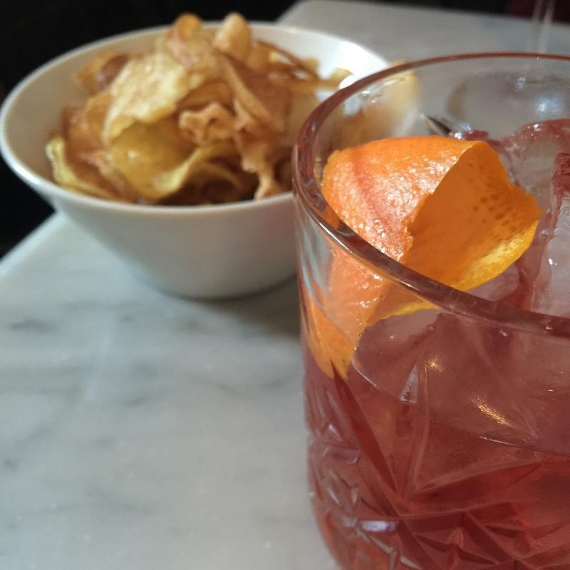 Auto-generated description: A glass of red Negroni cocktail with an orange peel garnish is placed on a table next to a bowl of potato chips.