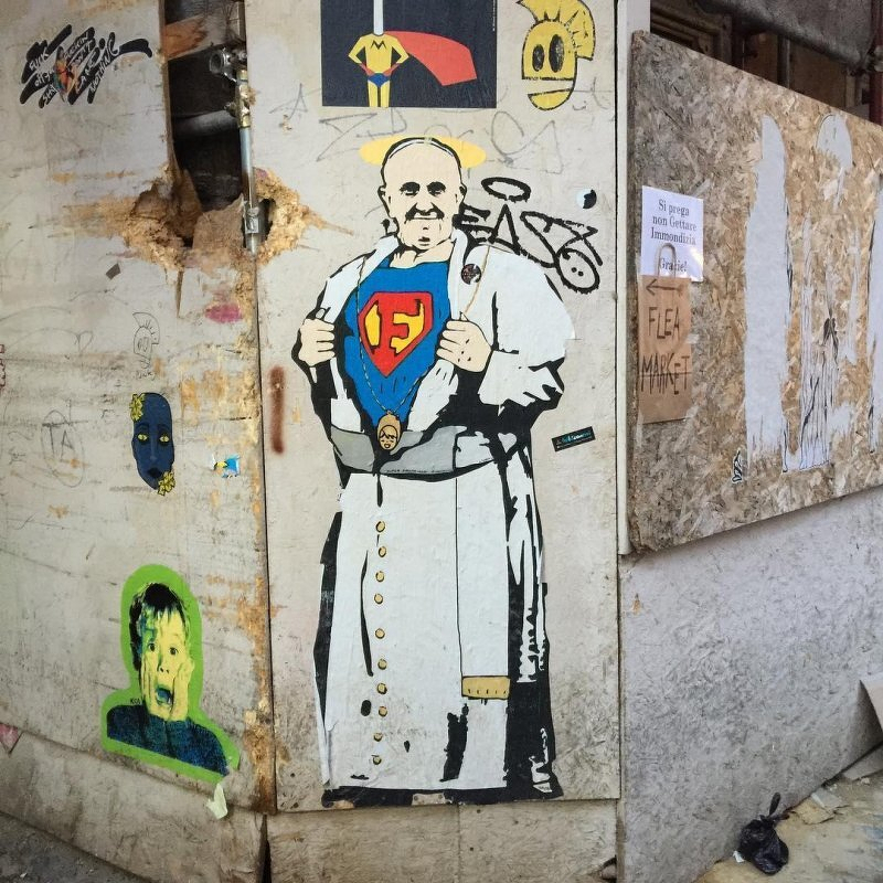 Auto-generated description: A colorful mural on a wall depicts Papa Francesco revealing a Superman-like costume under their robes, surrounded by various graffiti artwork and posters.