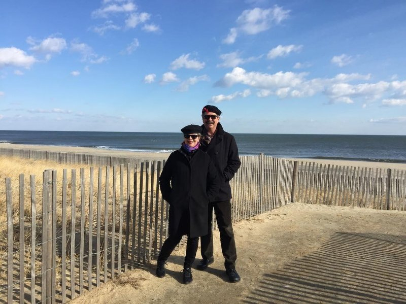 Auto-generated description: A couple wearing dark clothing and hats is standing on a sandy walkway by the beach, with a wooden fence and the ocean in the background.