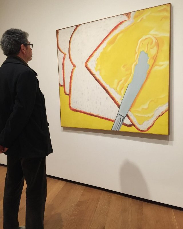 Auto-generated description: A person observes a large painting of bread being spread with butter in an art gallery setting.