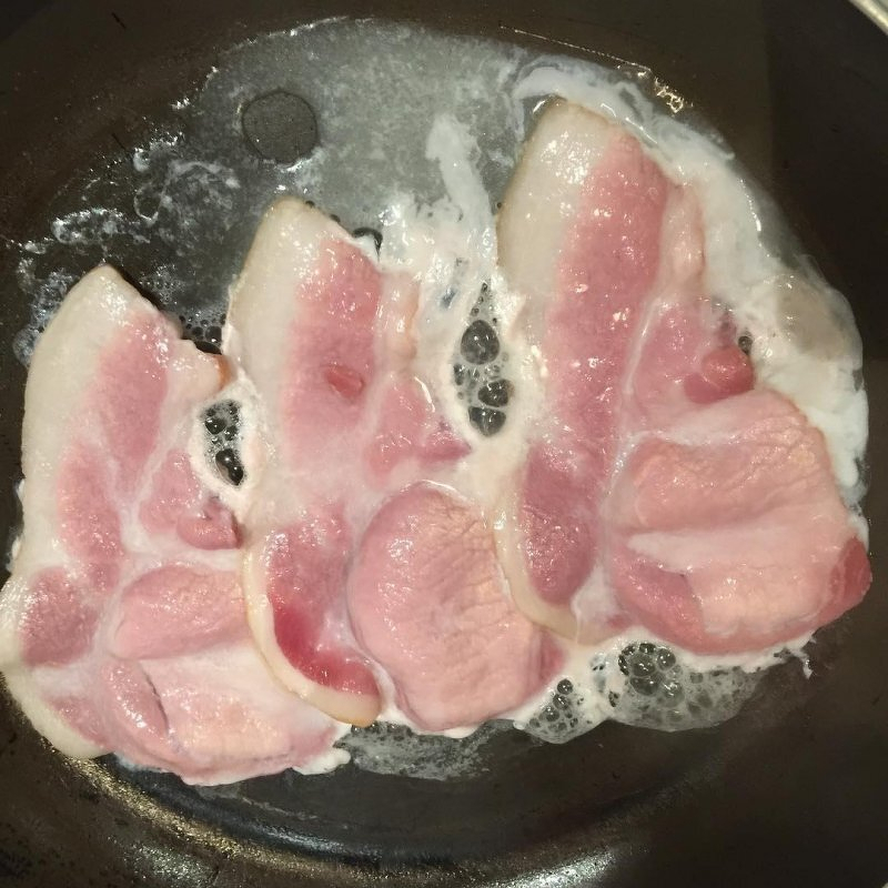 Auto-generated description: Sizzling in a pan, several strips of bacon are cooking, surrounded by bubbling fat.