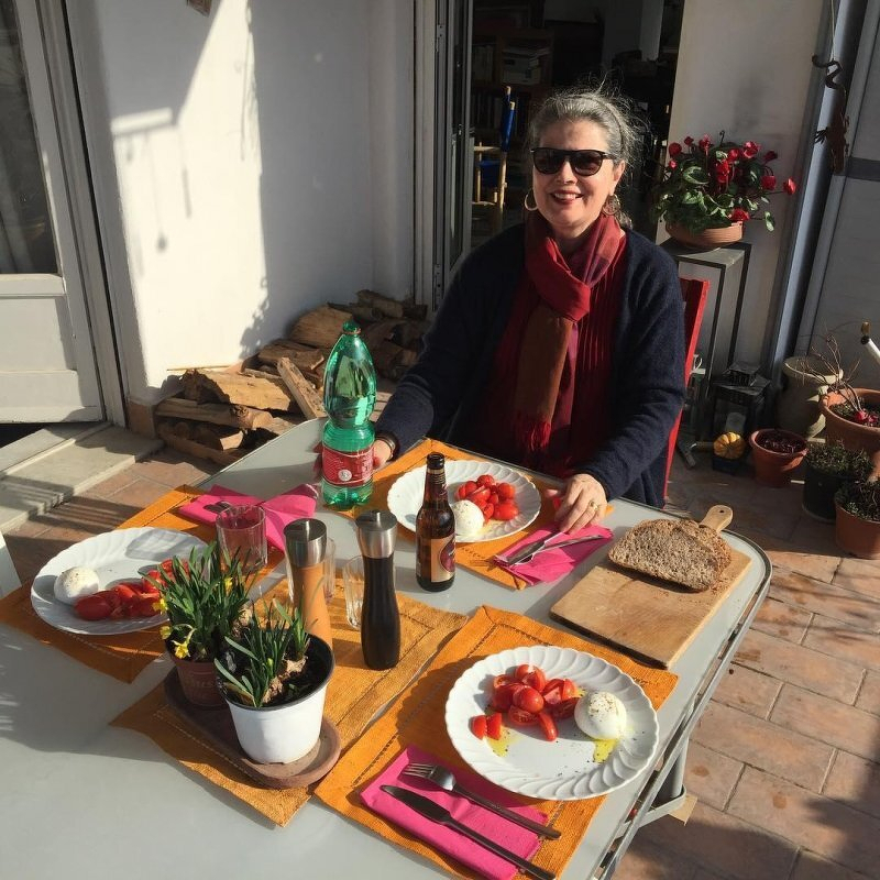 Auto-generated description: A woman is sitting outdoors at a table set with plates of food and drinks, enjoying a sunny day.