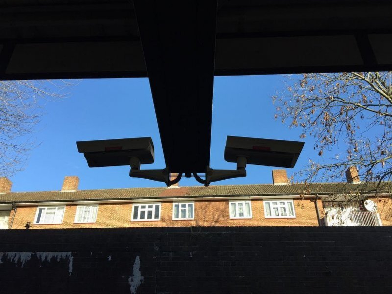 Two surveillancwe cameras against a blue sky with a row of houses behind.
