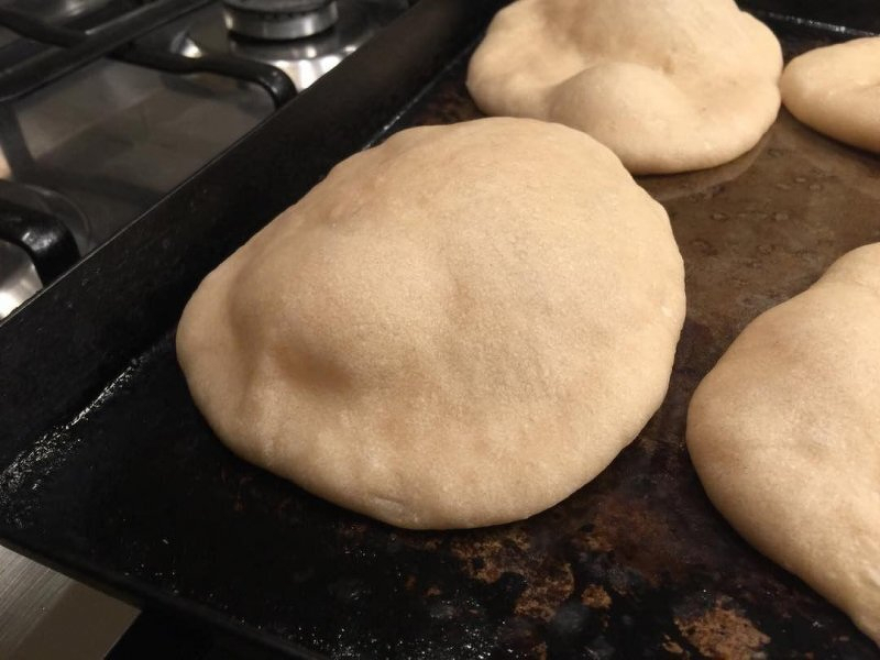 Auto-generated description: Several puffed-up pita breads are baking on a metal tray in the oven.