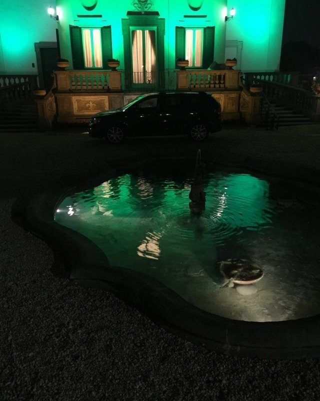 Auto-generated description: A small illuminated fountain and a parked car are in front of a grand building -- the Irish Embassy to the Holy See -- lit with green lights.