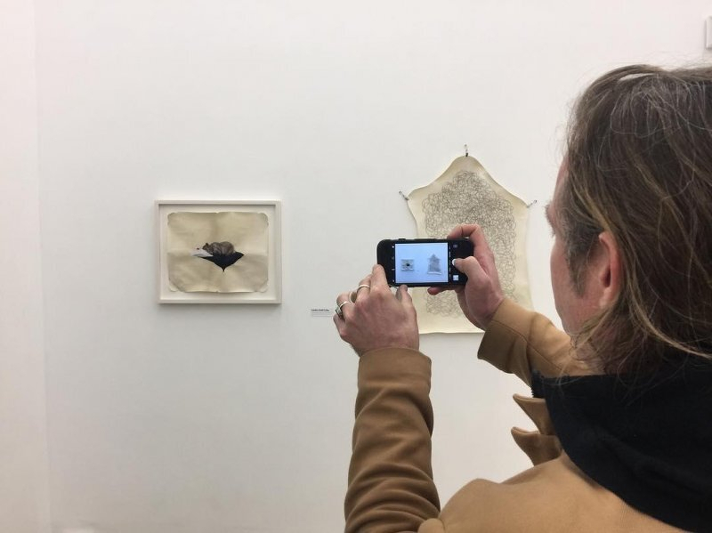 Auto-generated description: A person is photographing artwork on a gallery wall using a smartphone.