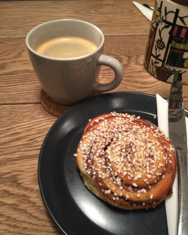 Auto-generated description: A cinnamon roll topped with pearl sugar is served on a black plate alongside a mug of coffee on a wooden table.