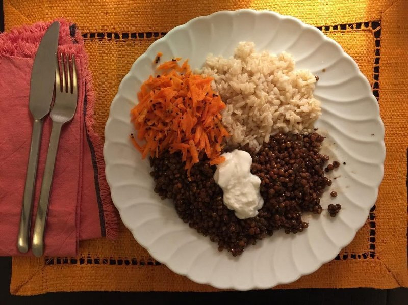 Auto-generated description: A plate holds portions of rice, grated carrots, lentils topped with a dollop of yoghurt, alongside a set of cutlery on a pink napkin.