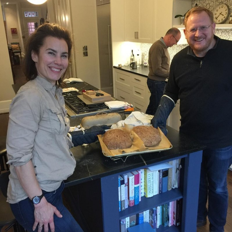 Auto-generated description: Two people are smiling in a kitchen while showing freshly baked loaves of bread on a counter, with another person working in the background.
