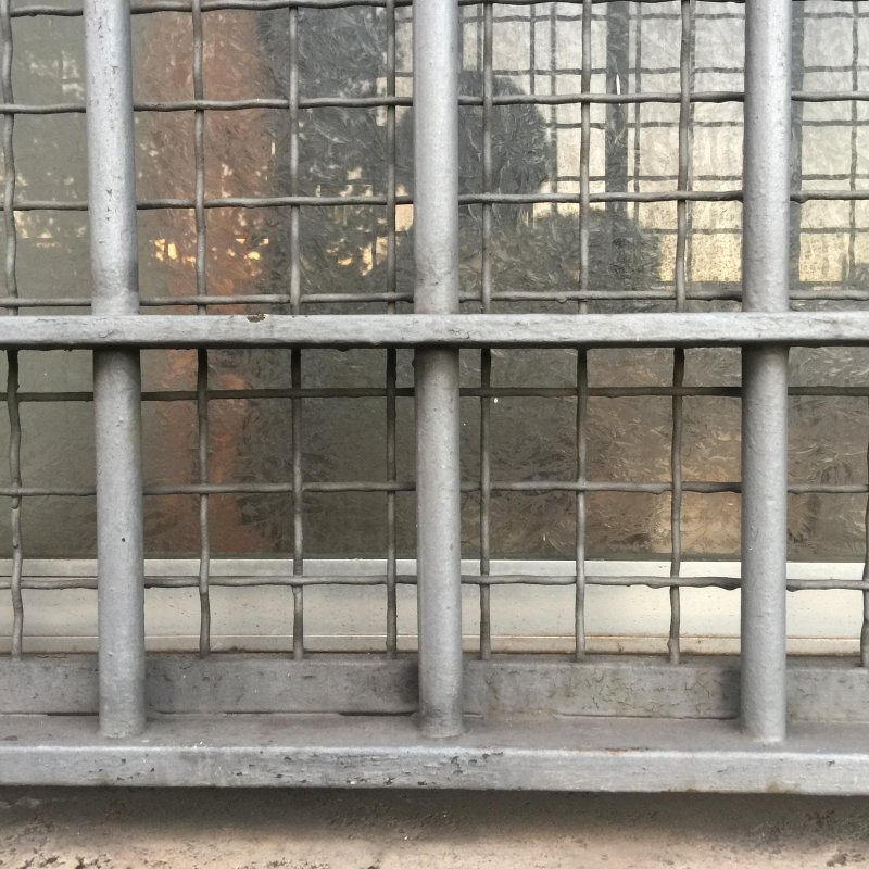 Auto-generated description: A metal grate with a grid pattern is mounted over a window, revealing a reflection in the glass.