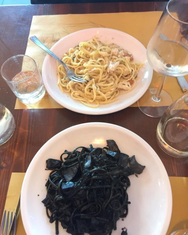  Two plates of pasta, one with carbonara sauce and the other with black squid ink sauce, are served alongside drinks on a wooden table.