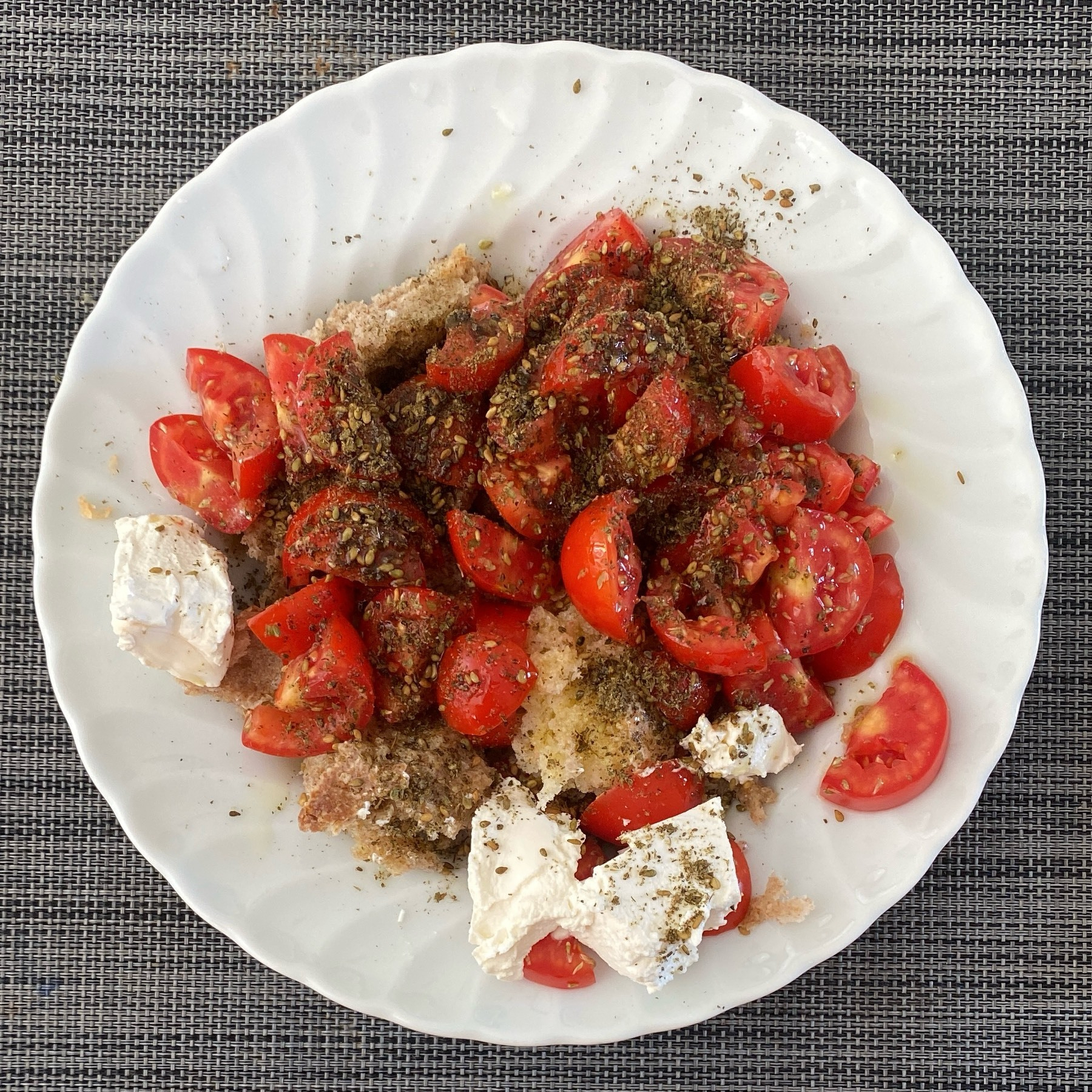 A white plate with quartered red tomatoes on top of stale bread with white ricotta cheese on the side. 