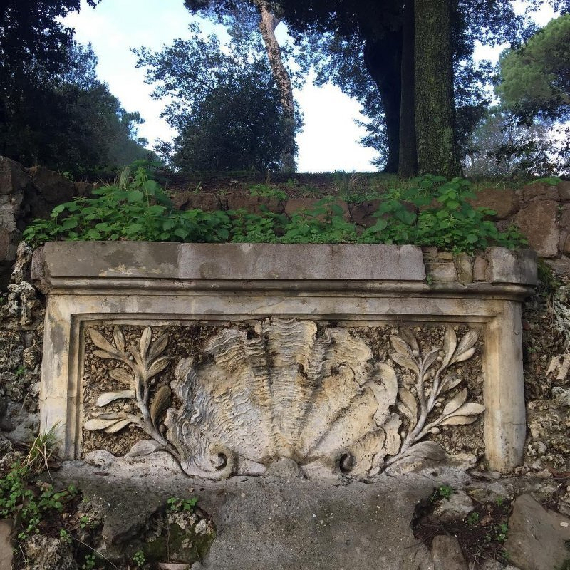 Auto-generated description: An ancient carved stone panel, featuring a large shell surrounded by botanical motifs, is set into a hillside with trees and greenery above.