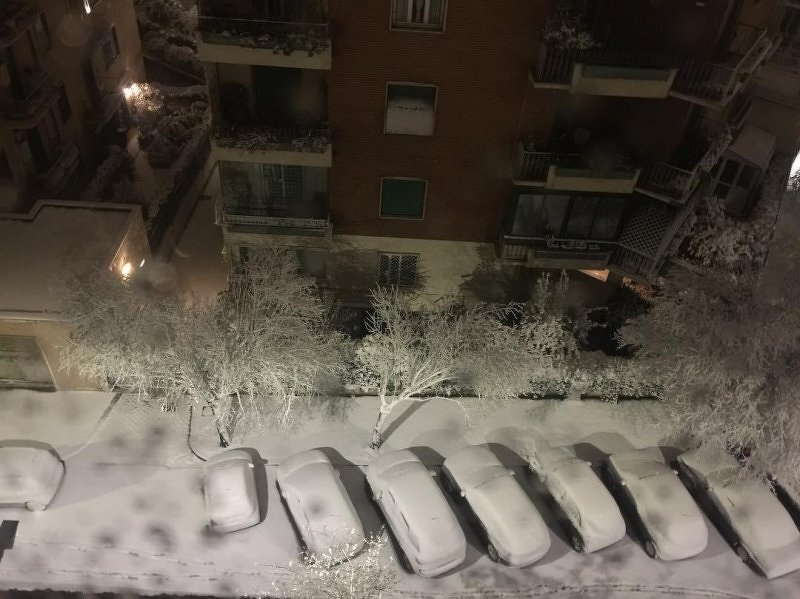 Auto-generated description: A snowy urban street is lined with cars and trees covered in snow, viewed from above.