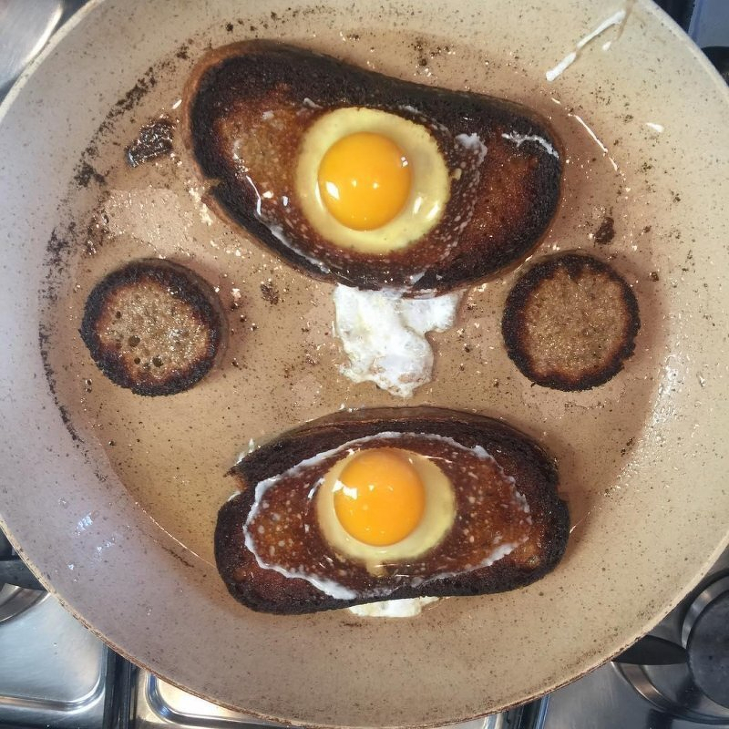 Auto-generated description: Two pieces of toast with fried eggs in the center are cooking in a pan alongside small, round pieces of bread, the remnants of the holes in which the eggs have been placed.