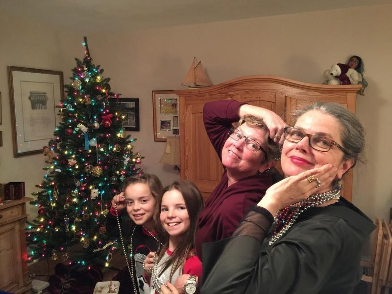 Auto-generated description: Four people are posing joyfully in front of a decorated Christmas tree, each holding or wearing a string of beads.