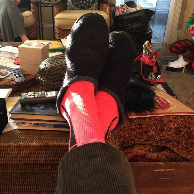 Auto-generated description: A person relaxing with bright pink socks and black slippers, feet propped up on a wicker table surrounded by books, remote controls, and household items.
