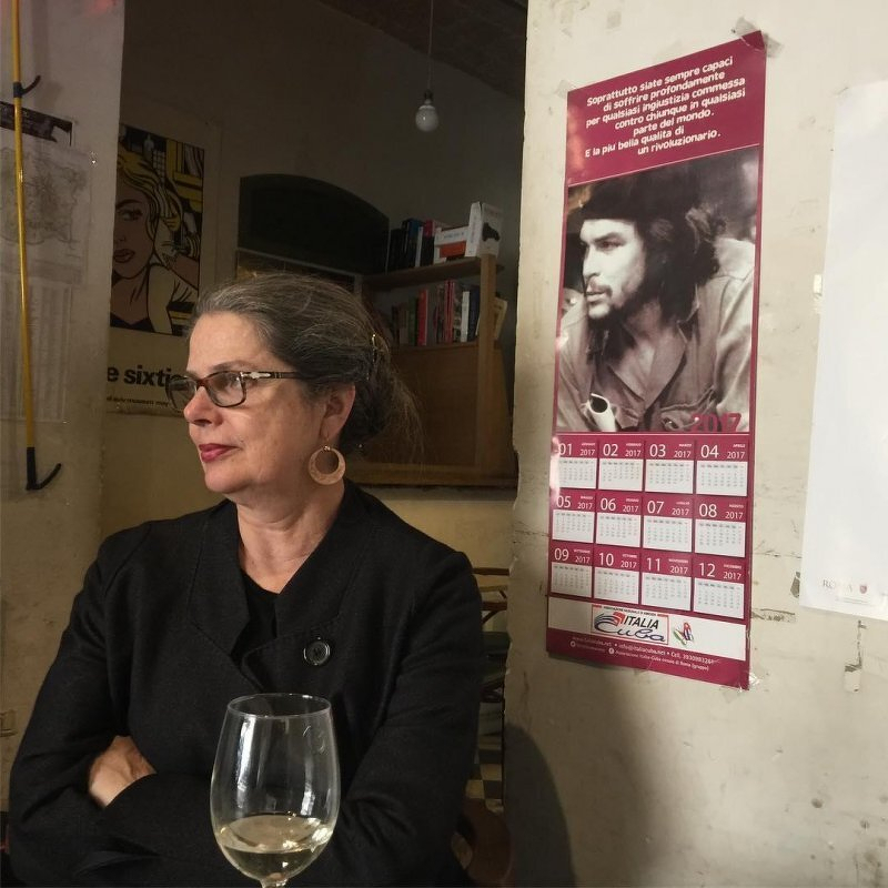 Auto-generated description: A woman with glasses sits in a room next to a calendar featuring Che Guevara and a glass of white wine.
