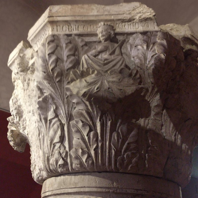 An intricately carved stone column capital features a figure sitting with flowing drapery amidst ornate leaf motifs